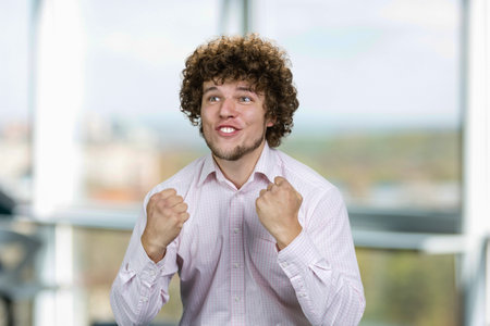 Portrait of happy excited young man with curly hair rejoicing success. Young football fan. Indoor window in the background.の写真素材