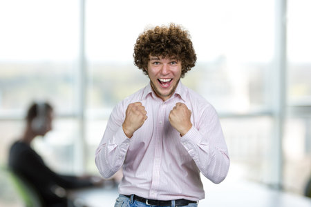 Portrait of happy excited young man with curly hair celebrates success. Indoor office interior in the background.の写真素材