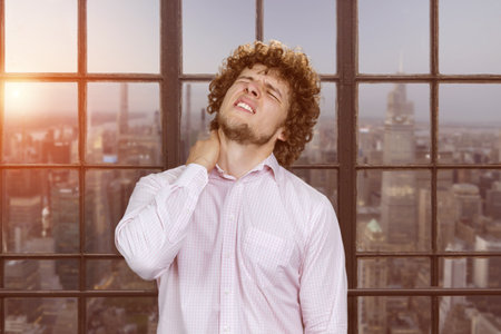 Portrait of a young caucasian man with curly hair having a neckache. Window with cityscape view in the background.の写真素材
