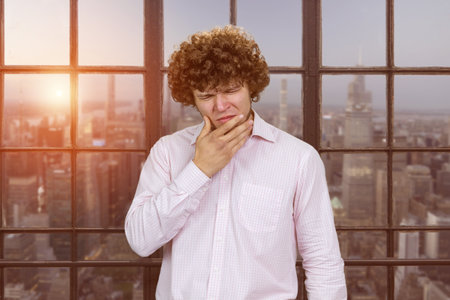Young man with curly hair touching his jaw because of toothache.の写真素材