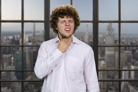 Young handsome man with curly hair suffers from neck pain. Indoor window with cityscape view in the background.の写真素材