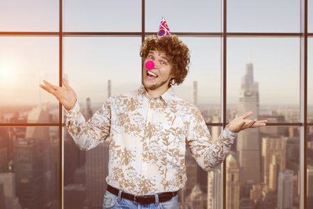 Portrait of a happy young man with clown nose. Indoor window with evening sunset view in the background.の写真素材