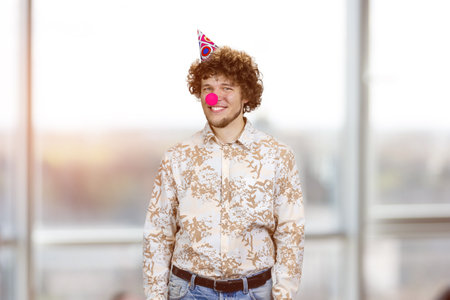 Portrait of happy young guy with clown nose. Indoor window in the background.の写真素材