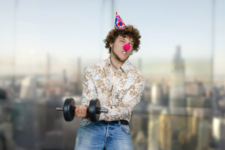 Portrait of a young clown making a gym exercise with dumbbell. Blurred cityscape in the background.の写真素材