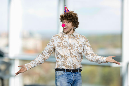 Portrait of happy young guy with clown nose and cone on his head. Indoor window in the background.の写真素材
