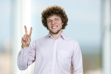 Portrait of a happy young man with curly hair shows victory gesture sign. Indoor window in the background.の写真素材