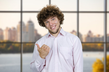 Portrait of happy cheerful young man with curly hair pointing back with his thumb. City river in the background.の写真素材