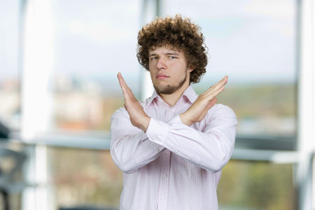 Portrait of a young man with curly hair cross his arms as sign of prohibition. Office window in the...