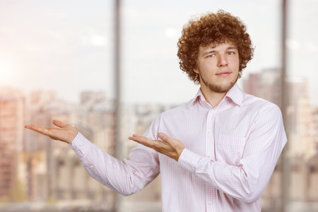 Portrait of a young man with curly hair advertising copy space with both hands. Cityscape in the background.の写真素材