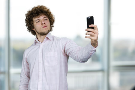 Young man with curly hair making a selfie with arrogant facial expression. Indoor window in the background.の写真素材