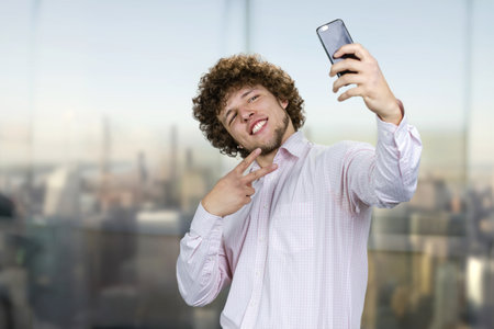Smiling young man with curly hair making a selfie with victory gesture sign. Urban cityscape in the background.の写真素材