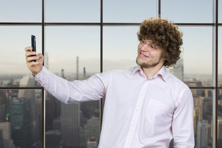 Young man with curly hair making a selfie and smiling with teeth. Indoor window with cityscape in the background.の写真素材
