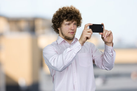 Portrait of a young tourist with curly hair making a photo on his smartphone camera. Blurred urban scape in the background.の写真素材