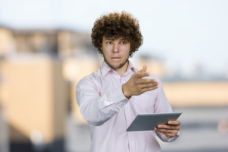 Portrait of a young caucasian worker with curly hair standing with tablet pc device pointing at something. Blurred urban scape in the background.の写真素材