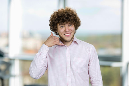 Handsome young man with curly hair shows call me gesture. Indoor window in the background.の写真素材