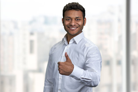 Smiling indian businessman looking at camera and giving thumb up gesture. Young smart man gesturing thumb up on blurred background.の写真素材