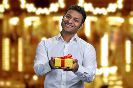Successful Indian businessman offering gift box on golden bokeh background. Present for Christmas holiday.の写真素材
