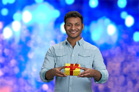 Attractive smiling young man handing gift box to camera. Blue bokeh lights background. Christmas present delivery in time.の写真素材