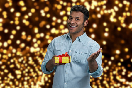 Delighted young man showing gift box and looking at camera. Festive bokeh background. Best Christmas gift shop.の写真素材
