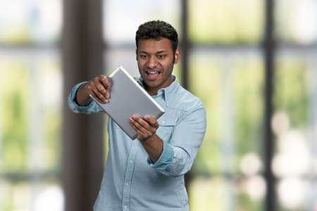 Energetic young man wearing blue shirt playing game on computer tablet. Interior blur background.の写真素材