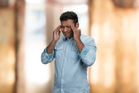 Stressed indian man massaging his temples because of terrible migraine. Exhausted young man standing on blur interior background.の写真素材