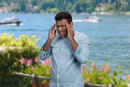 Stressed young man suffering from terrible headache standing outdoors. Man with headache on sea background.の写真素材