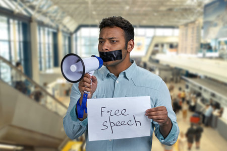 Young man with taped mouth trying to speak into megaphone while standing on escalator at airport. Censored man holding card with inscription Free speech while standing on the sliding walkway in the airport.の写真素材