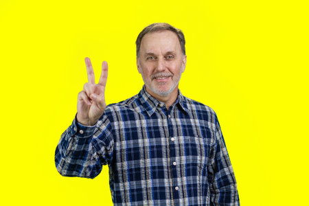 Smiling senior man showing victory gesture sign. Isolated on yellow.
