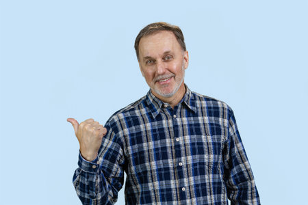 Portrait of smiling mature man pointing back with his thumb up. Isolated on pale blue.の写真素材