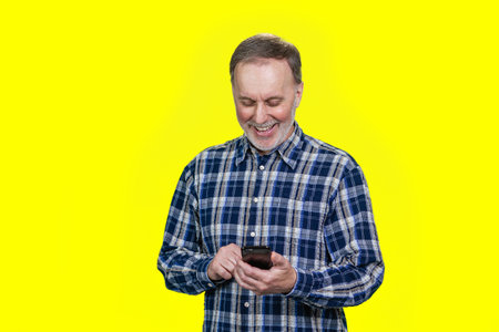 Portrait of aged man is holding his smartphone and typing a message. Isolated on yellow.