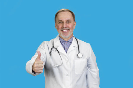 Smiling male aged senior doctor wearing stethoscope giving his thumb up. Isolated in the blue background.の写真素材