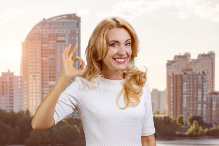 Portrait of a happy mature blond woman showing ok gesture sign. Residential area in the background.の写真素材