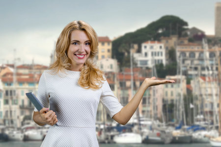 Portrait of cheerful blonde woman with tablet device shows copy space in front of a yacht docks. Vacation and tourism.の写真素材