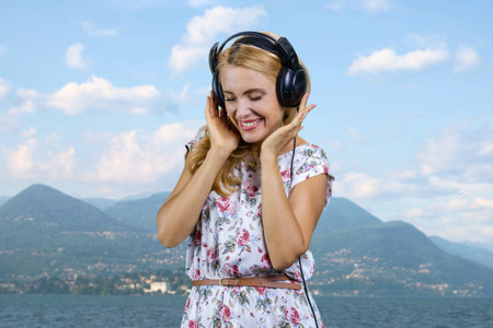 Mature blonde woman with headphones enjoying music outdoors. Mountain landscape in the background.の写真素材