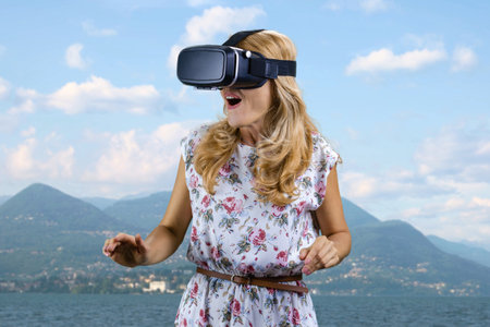 Portrait of happy amazed mature woman in virtual reality headset standing outdoors. Mountain...