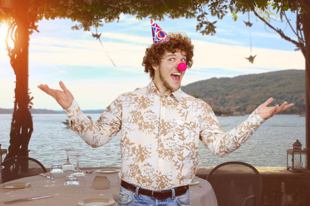Portrait of a young clown with curly hair spreads his arms. Sea landscape in the background.