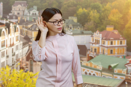 Asian woman in glasses is overhearing something. Outdoor cityscape background.