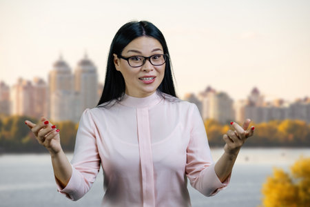 Young asian woman in glasses is standing outdoors and gesturing. River scape background.の写真素材