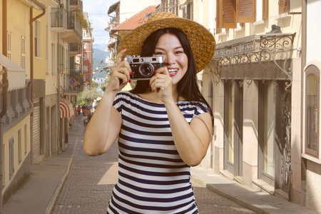 Portrait of a cheerful asian woman in straw hat holding vintage photo camera. European street...