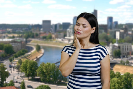 Young woman touching her cheek because of toothache. Urban cityscape background.の写真素材