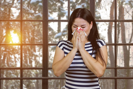 Asian woman with runny nose standing indoors. Sneezing brunette on checkered windows background with winter evening landscape.の写真素材