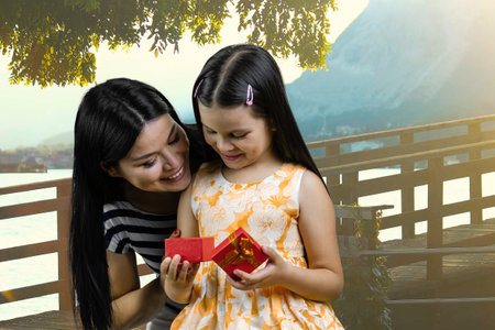 Brunette mother opens a red gift box to her little daughter. Sunny landscape background.の写真素材