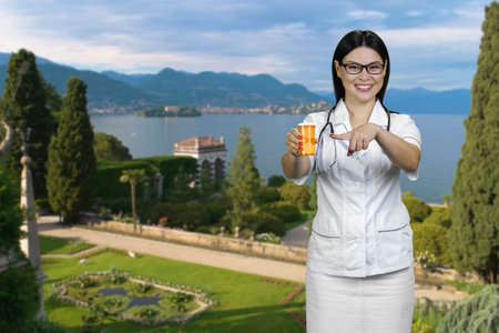 Young cheerful female doctor standing outdoors pointing at orange container of pills. Summer resort landscape background.の写真素材
