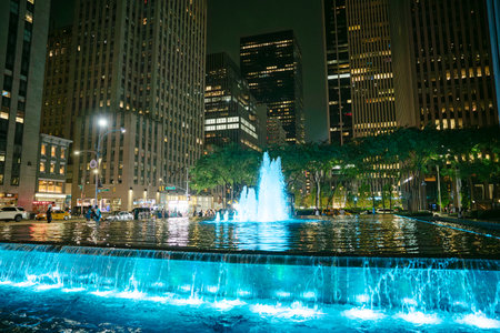 In Manhattan, a stunning fountain glows with blue light at night, surrounded by skyscrapers. People stroll along the water, enjoying the vibrant atmosphere of the city.の写真素材