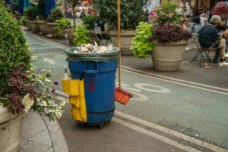 A blue trash bin with a broom and cleaning supplies stands on a street in New York. Large planters and people are seen in the background of this urban setting.の写真素材