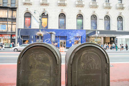 Two classic green U.S. Mailboxes stand on a red-painted curb in Manhattan. In the background, pedestrians walk past a commercial building with large windows and banners.の写真素材