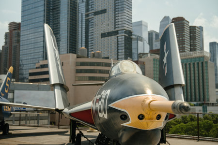 A restored U.S. Navy fighter jet with bold markings is displayed on an outdoor deck in front of tall city buildings. The image shows a close-up view of the cockpit, nose, and folded wings of the aircraft.の写真素材