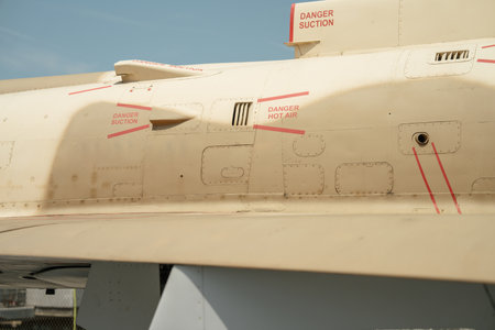 Close-up view of a beige military aircraft showing red warning labels and vent systems. Markings...