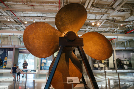 A large bronze ship propeller is mounted on a metal support inside a maritime museum. Museum visitors, including children, observe the impressive exhibit in a well-lit space.の写真素材