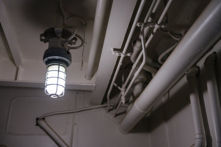 A glowing industrial-style ceiling lamp is mounted among painted metal pipes and conduit lines. The...